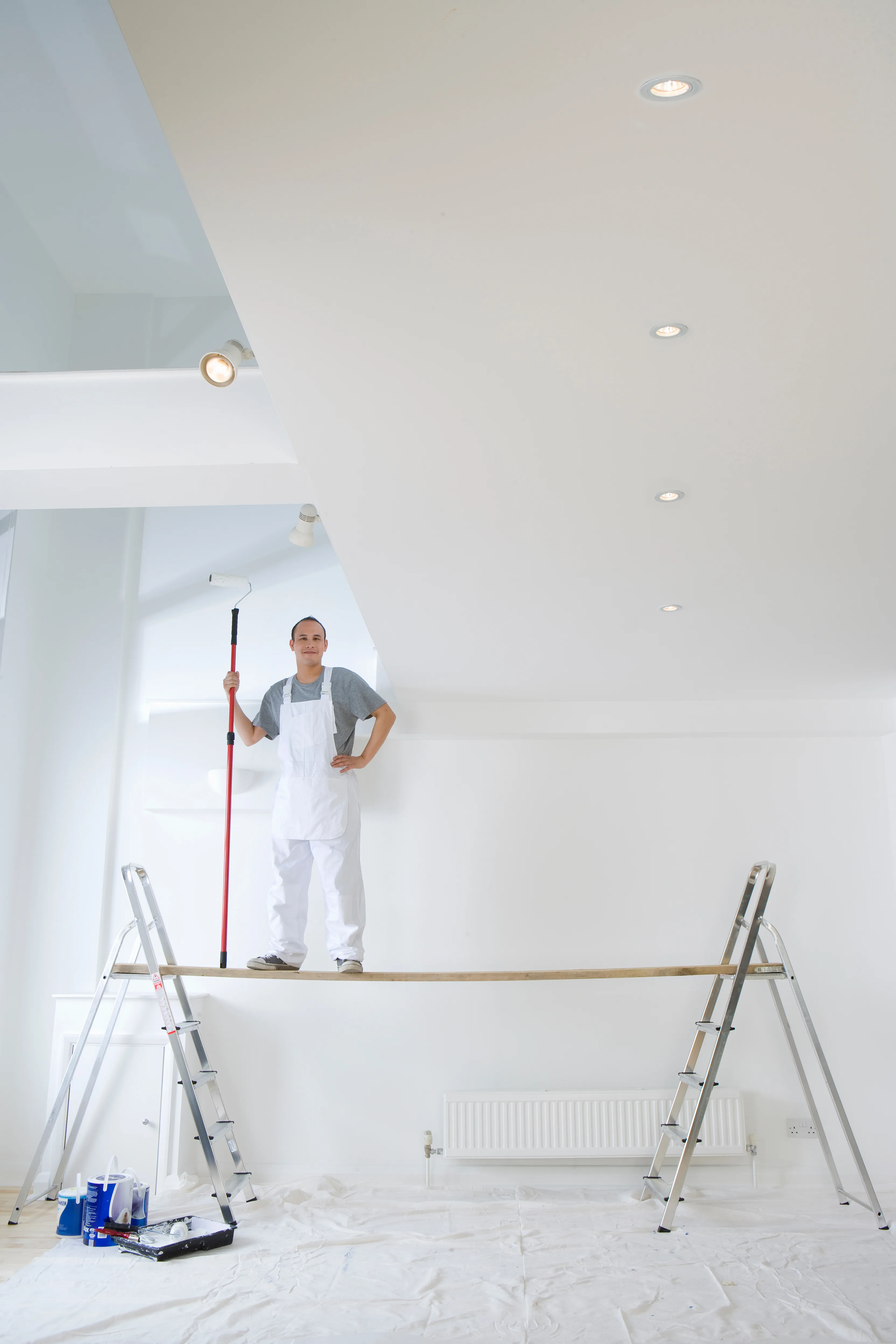 A painter in white overalls is applying paint to a ceiling while standing on a scaffold