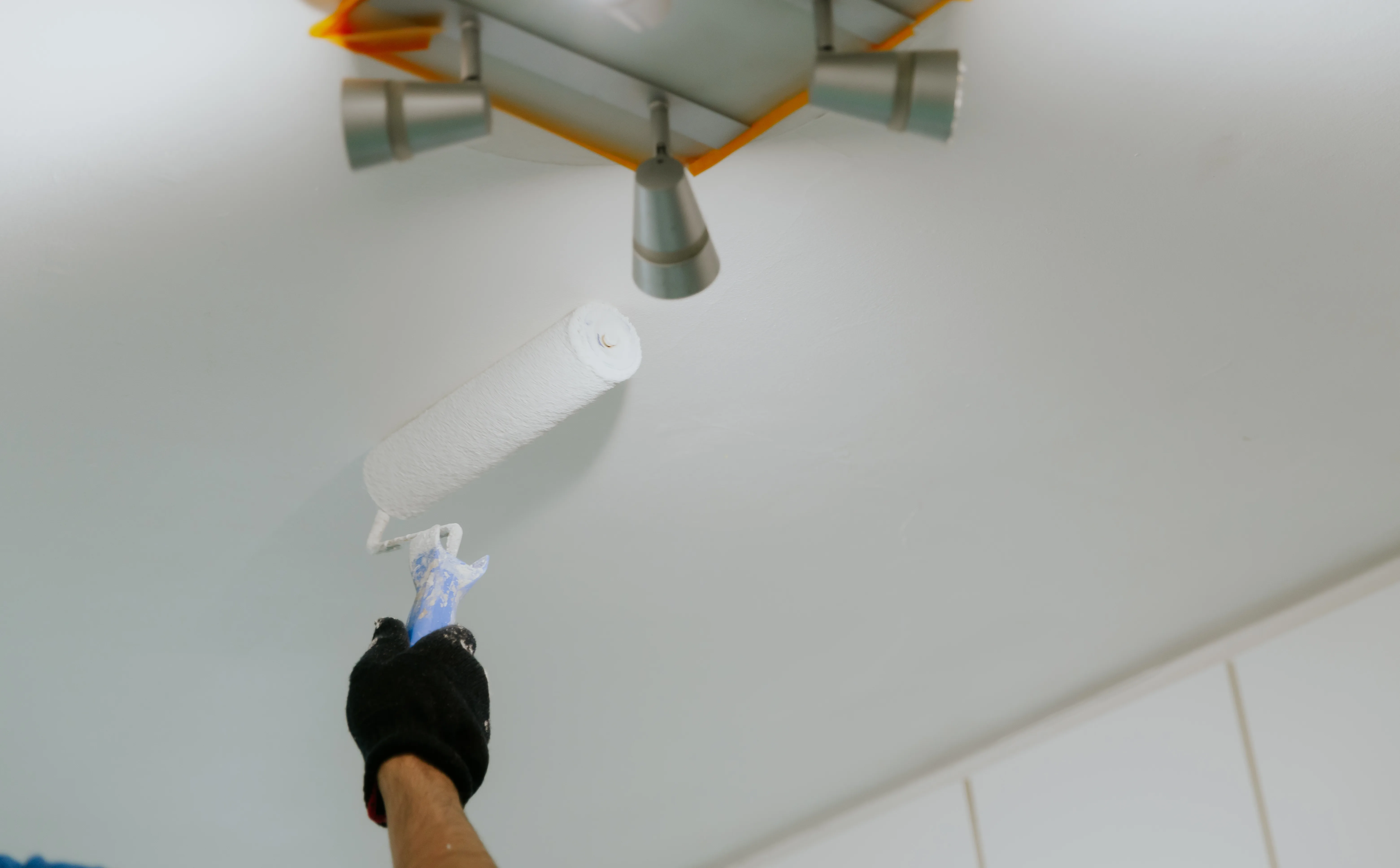 A hand painting a ceiling with a roller while a modern light fixture is visible