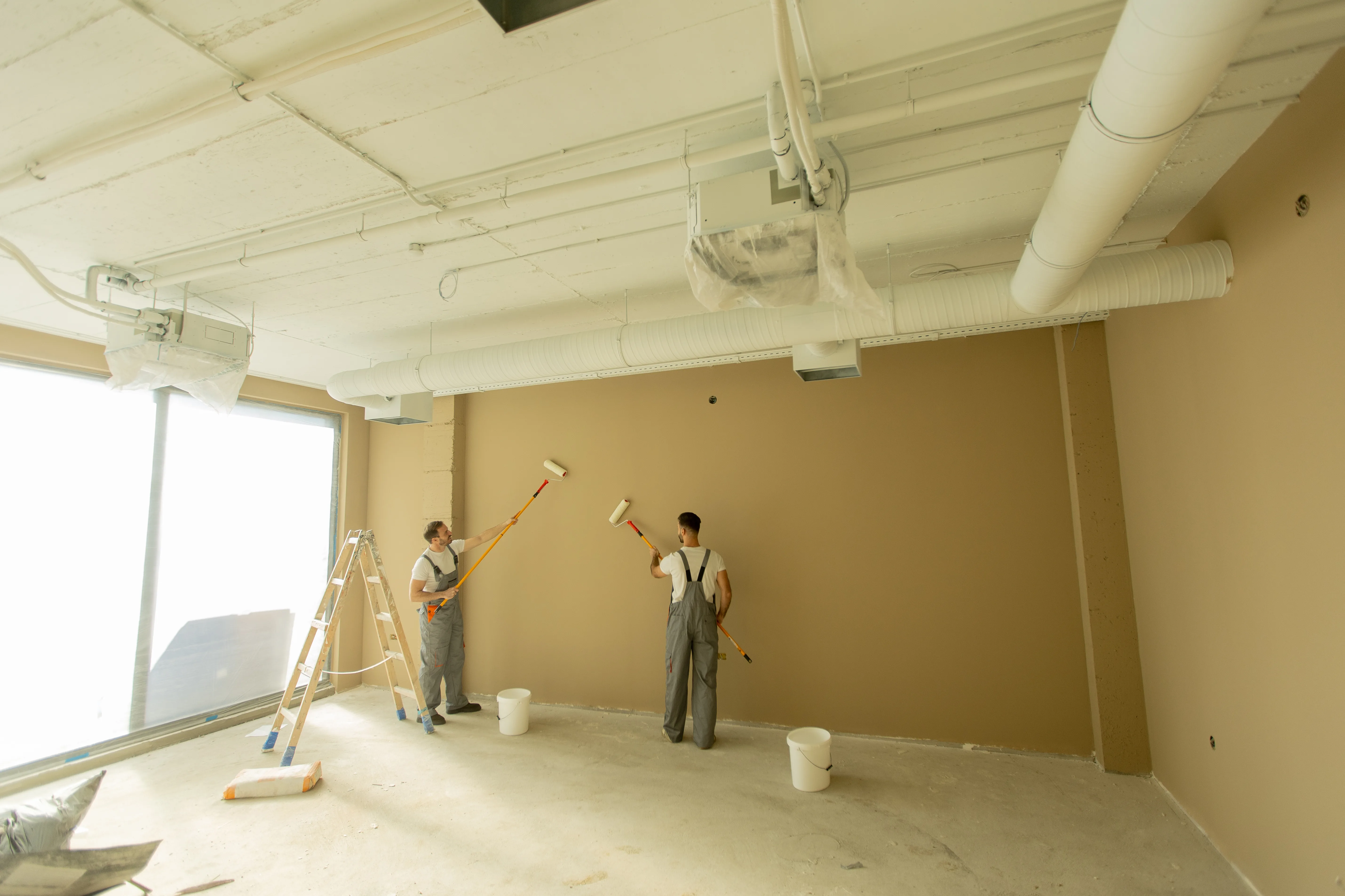Two painters applying wall paint in a modern interior space with a light brown wall
