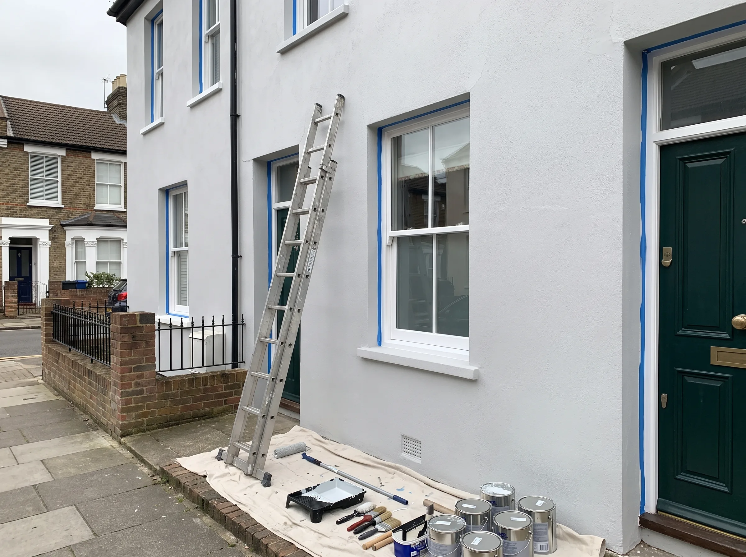 Exterior house painting in progress, with a ladder, paint cans, and prepared surfaces visible