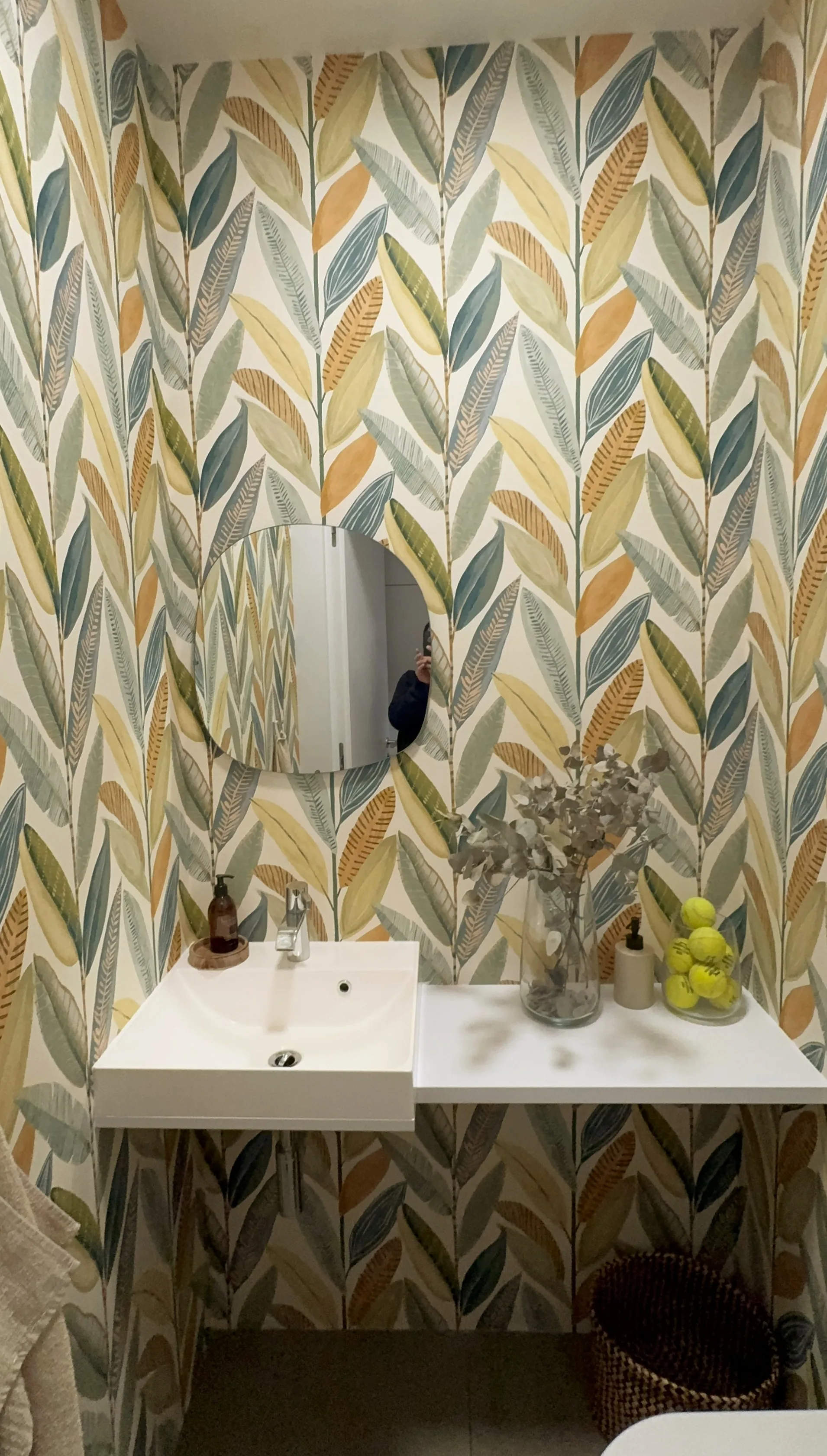 A bathroom interior featuring leaf-patterned wallpaper, a round mirror, and a modern sink with decorative items