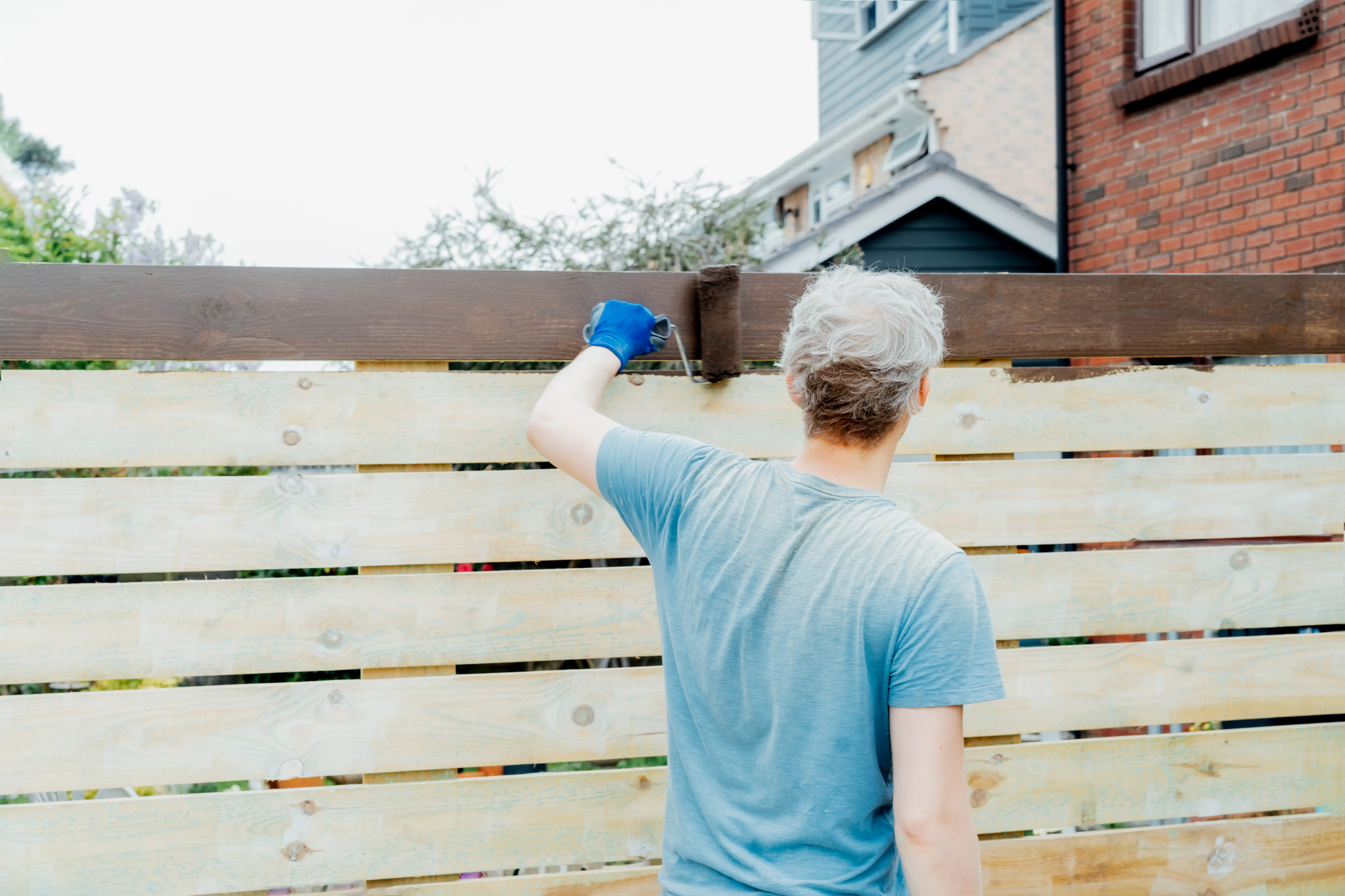 A person painting a wooden fence with a brush in a residential area