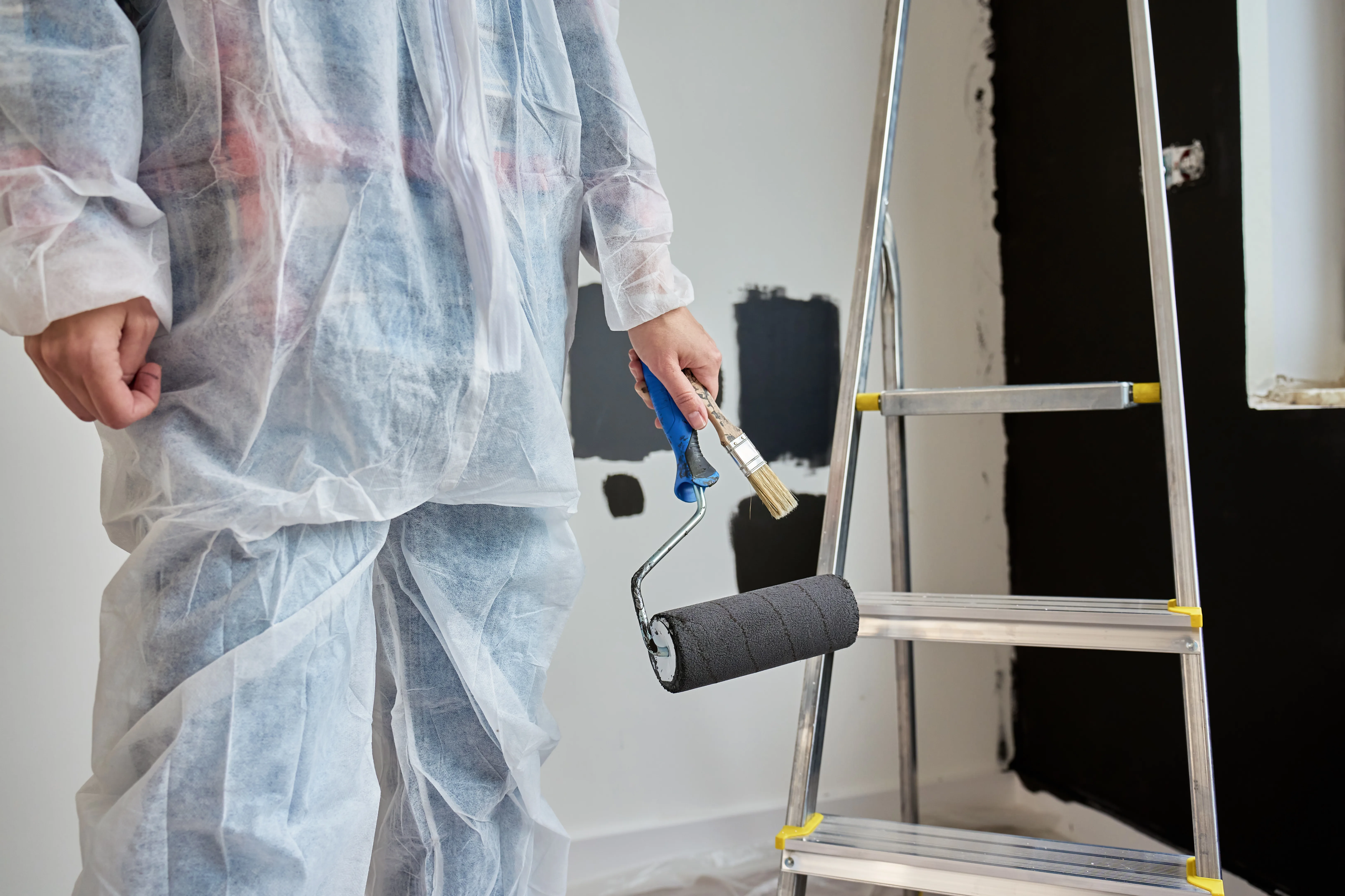 Person in protective suit holds a paint roller and stands by a ladder in a room being repainted