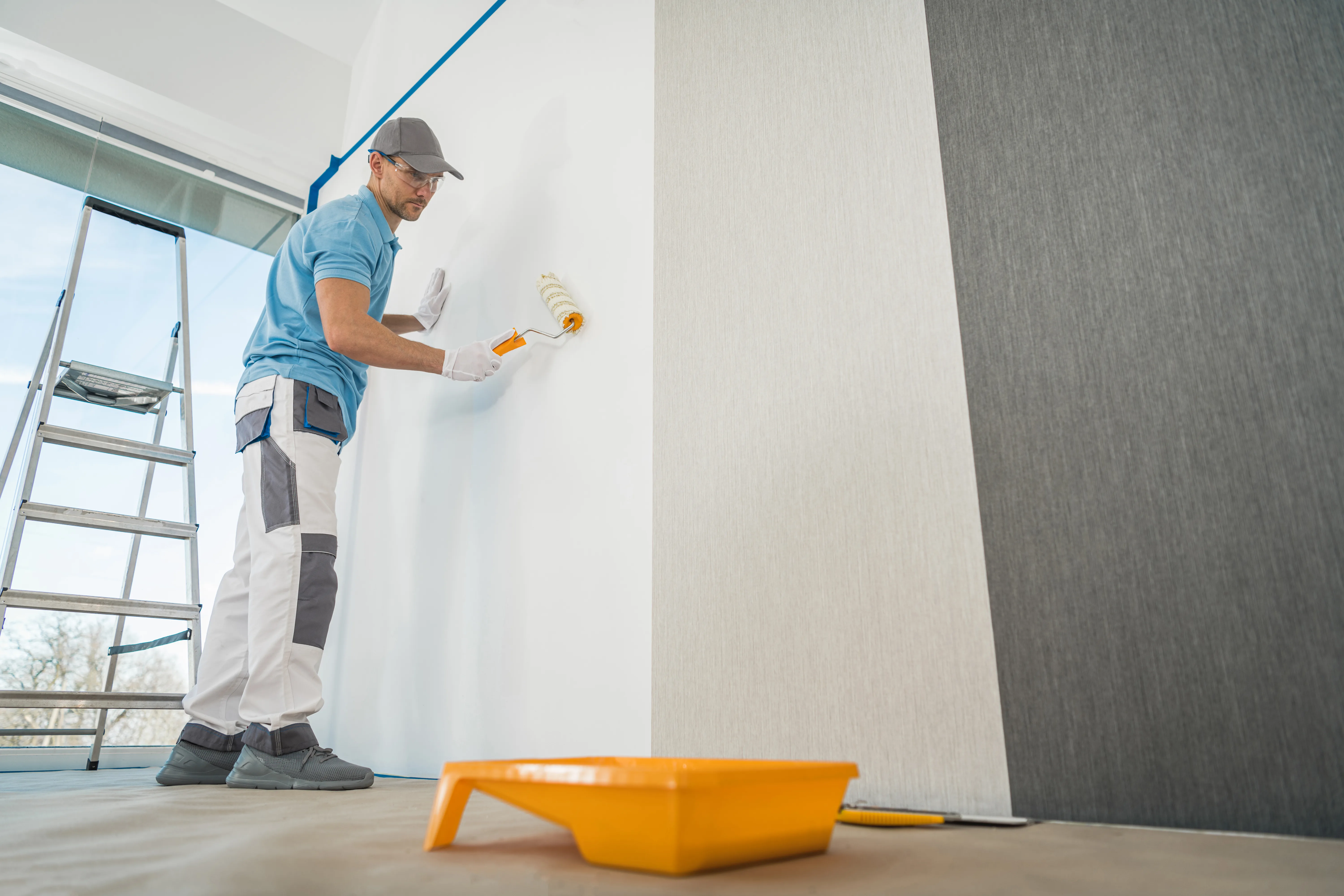 A painter applying paint to a wall using a roller, with a ladder and paint tray nearby