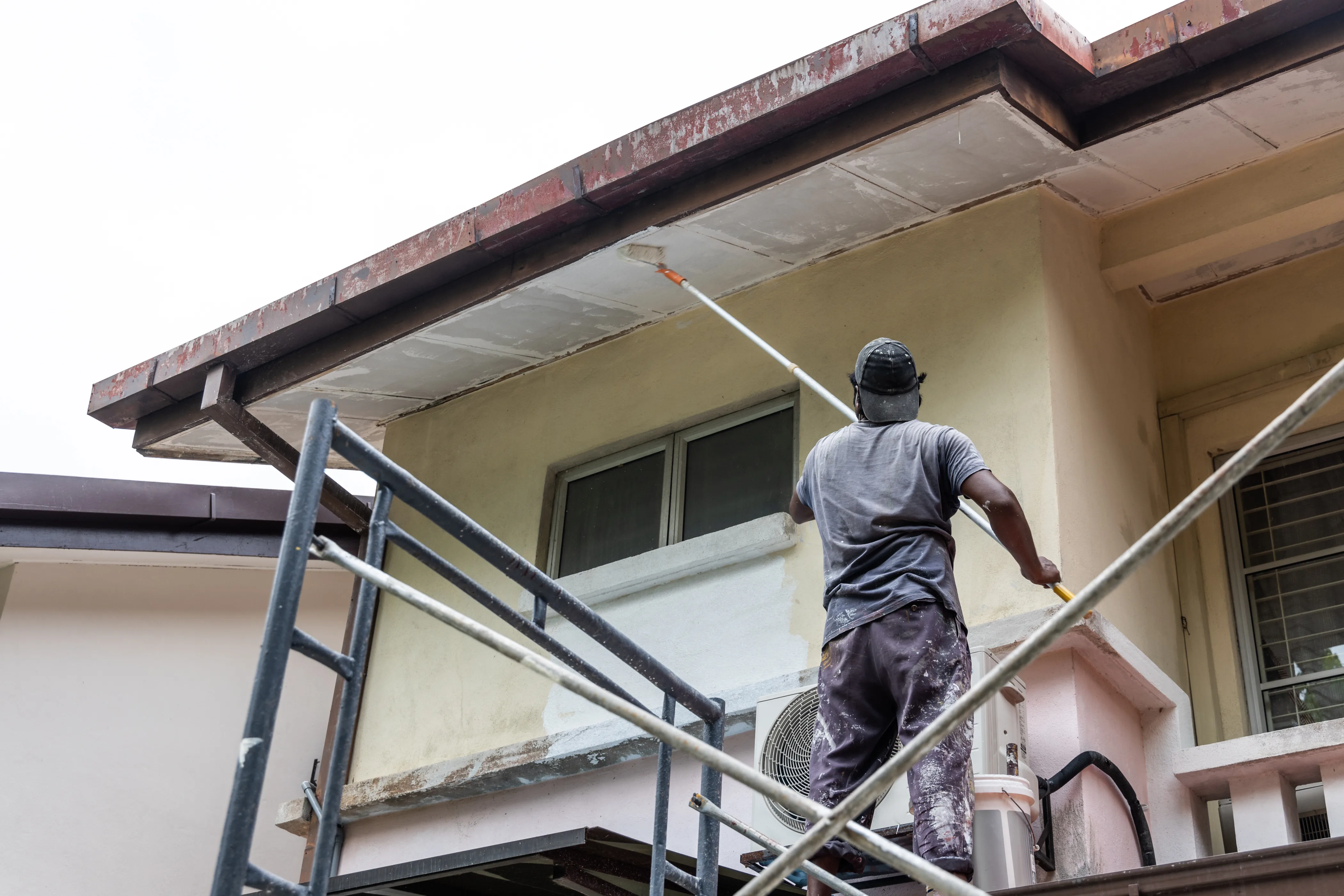 A painter applying exterior wall paint to a house using a roller on scaffolding