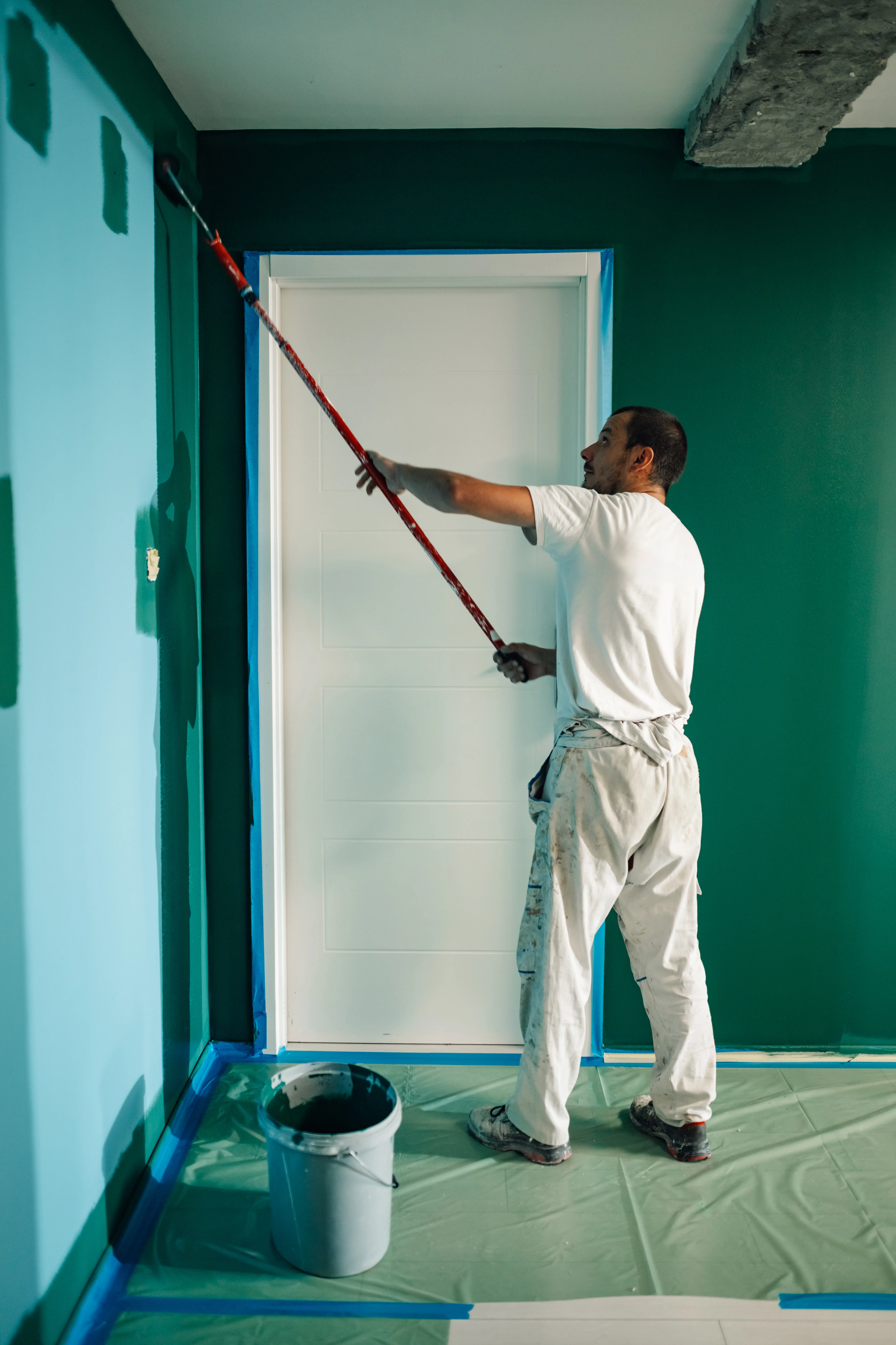 A painter applying green paint on a wall with a roller in a well-prepared room