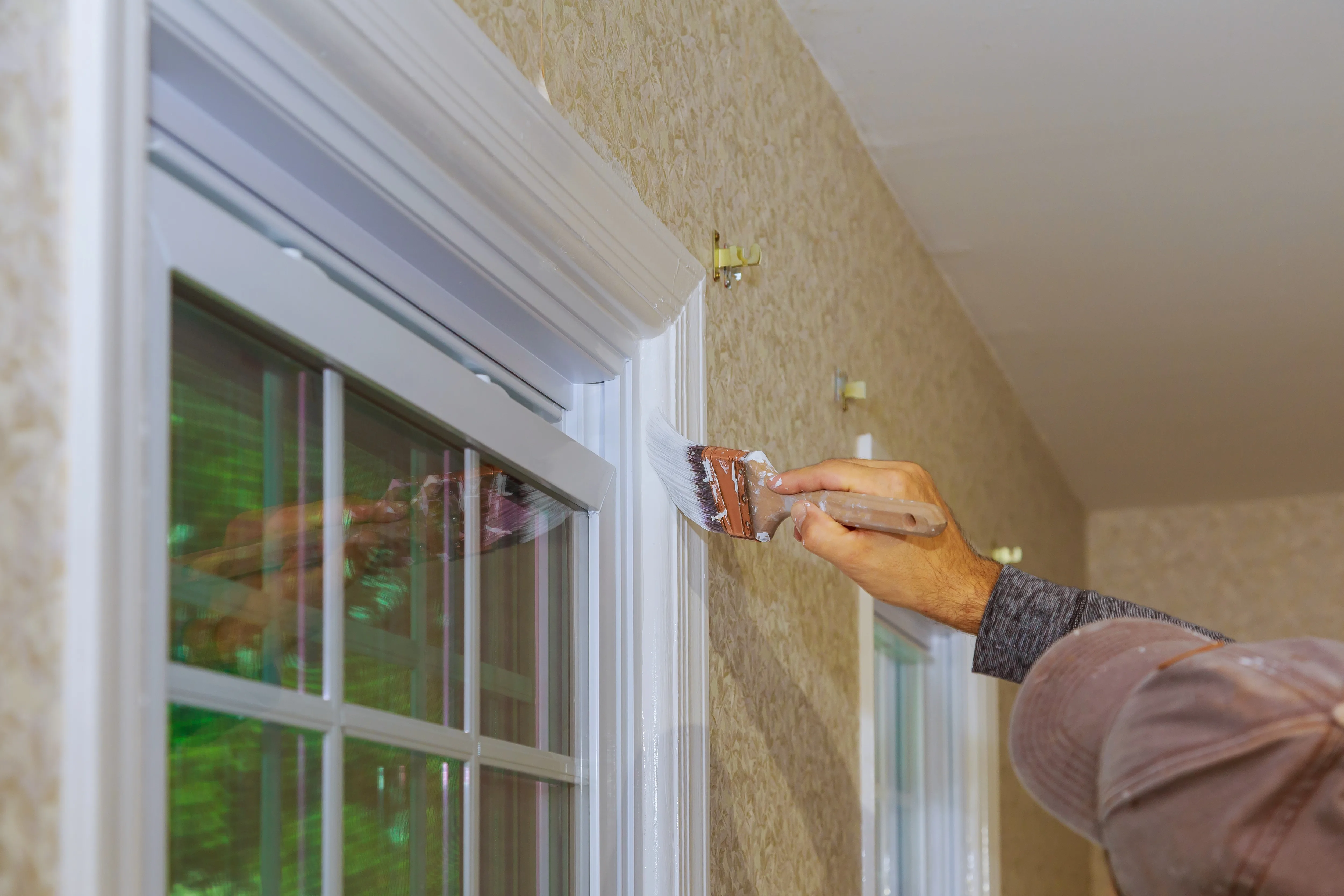 A painter using a brush to apply white paint to interior woodwork near a window