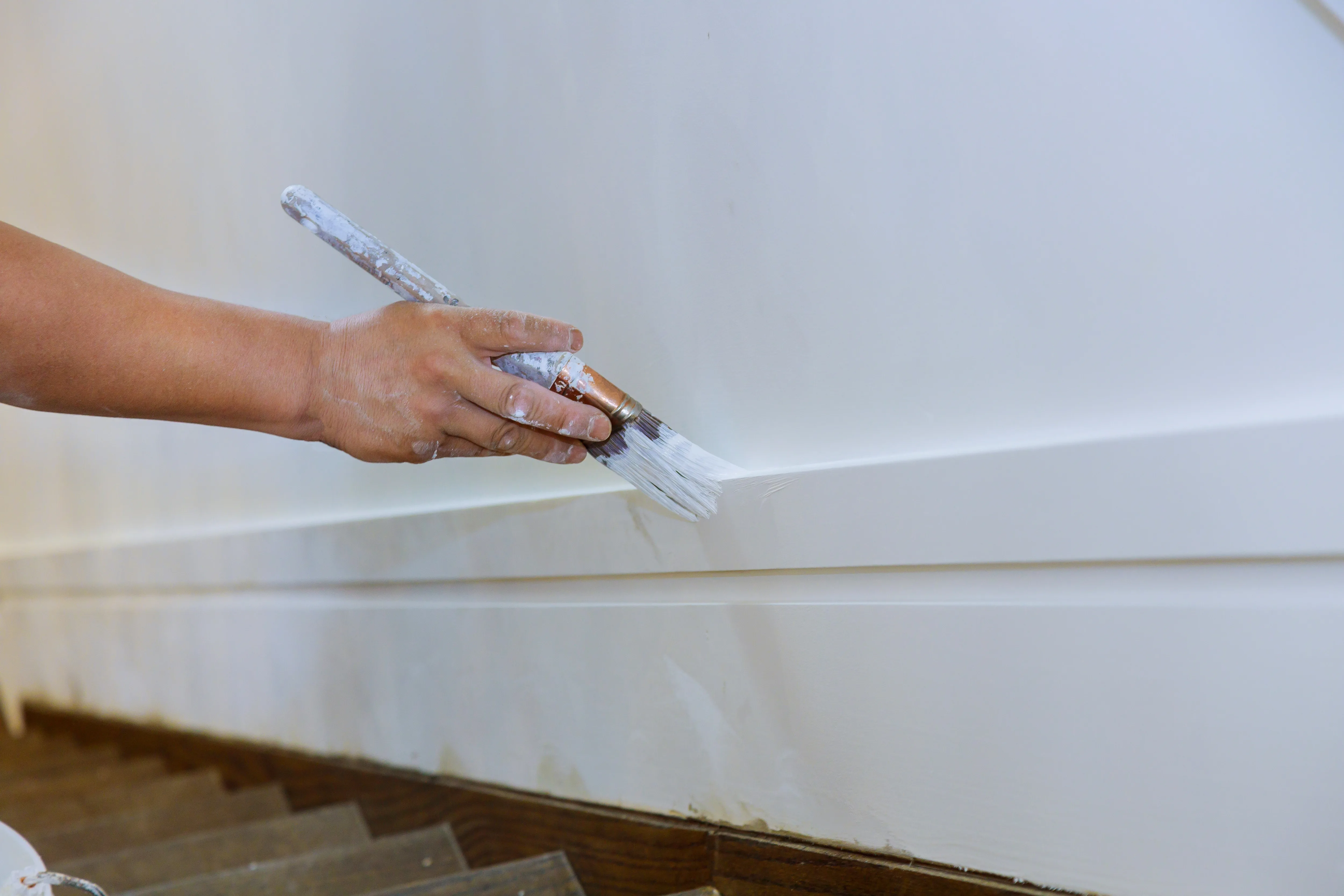 Close-up of a painter's hand applying white paint on trim near stairs