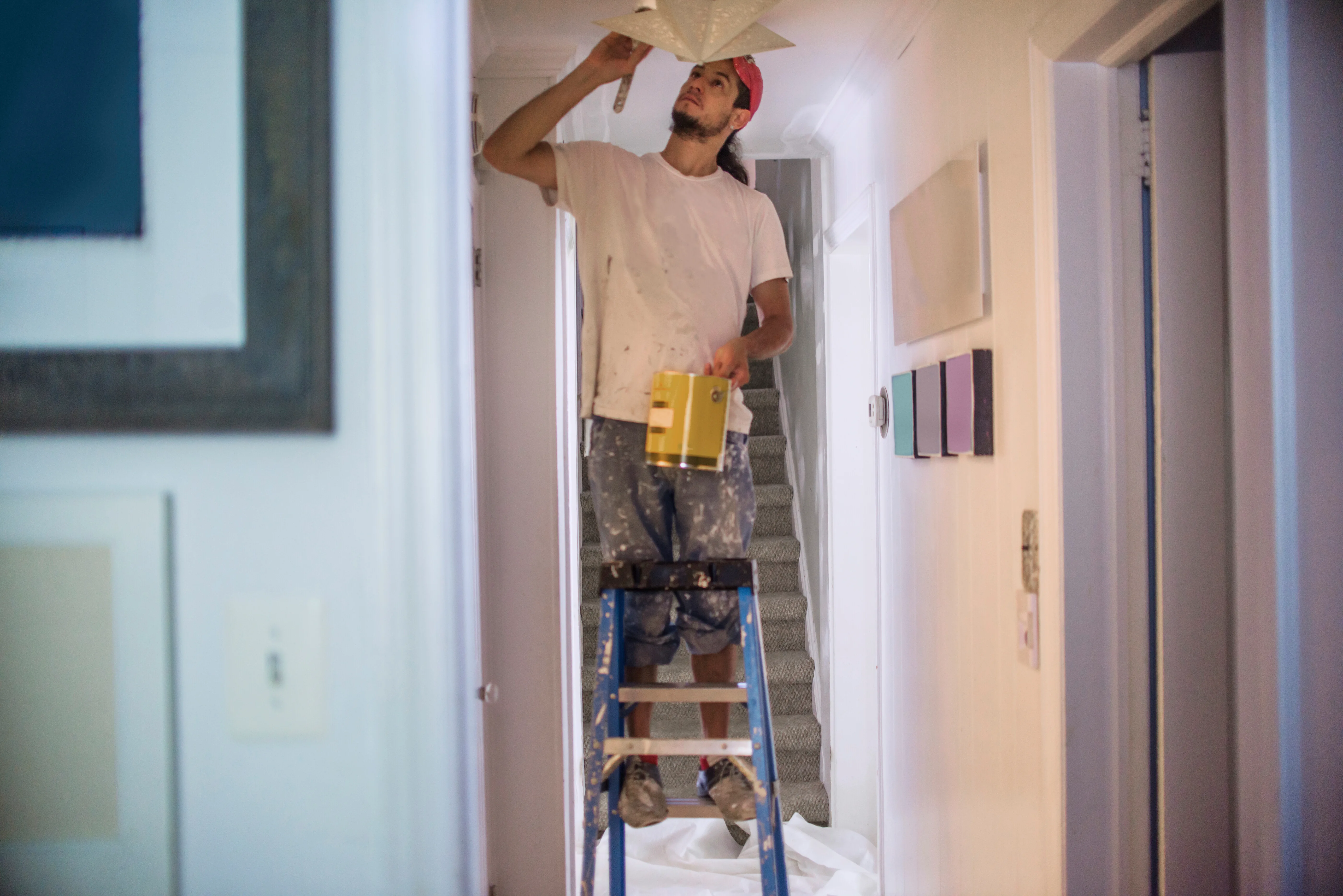 A painter on a ladder adjusting a ceiling light fixture in a hallway