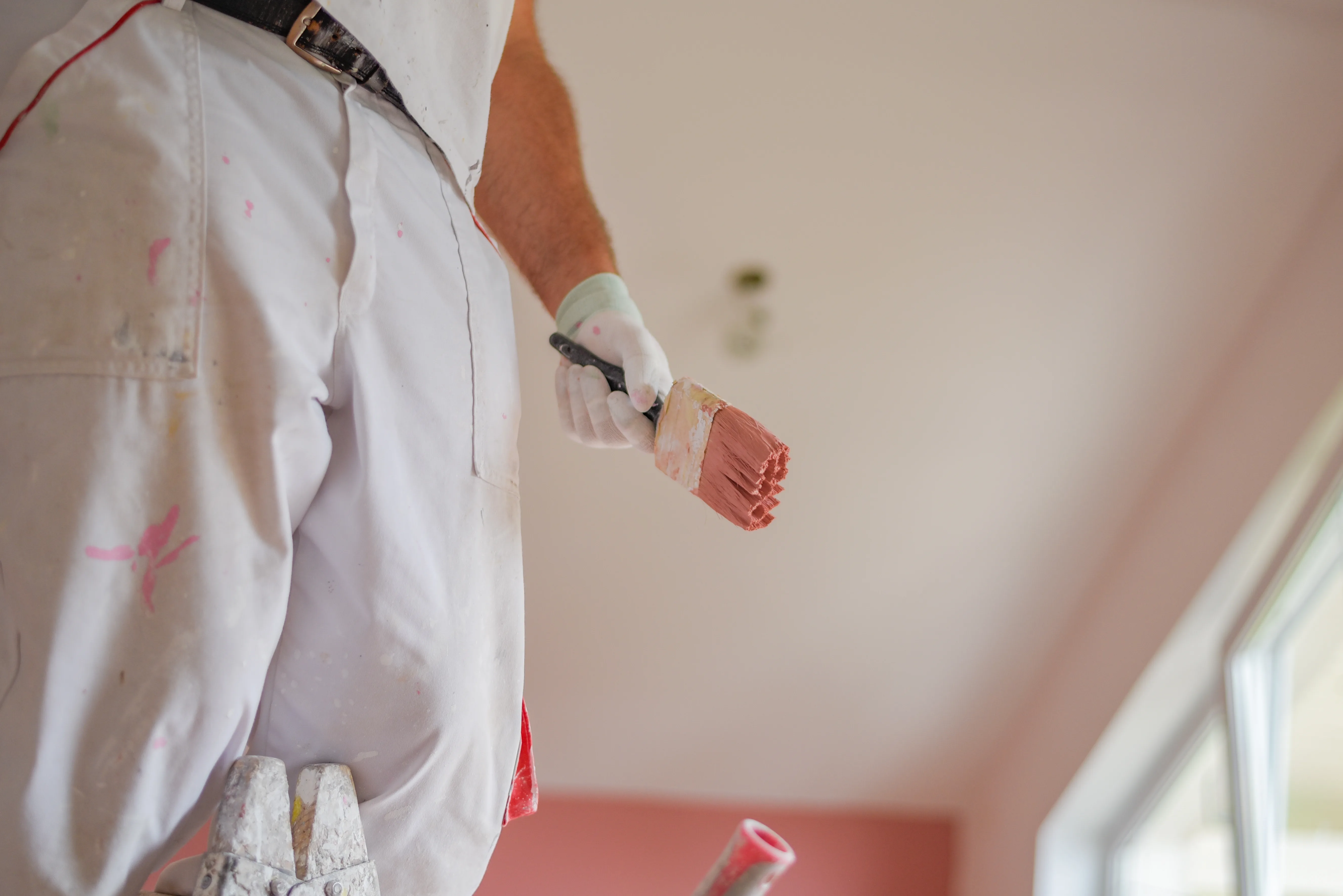 A painter holding a paintbrush while standing on a ladder, preparing to paint a ceiling in a light pink room