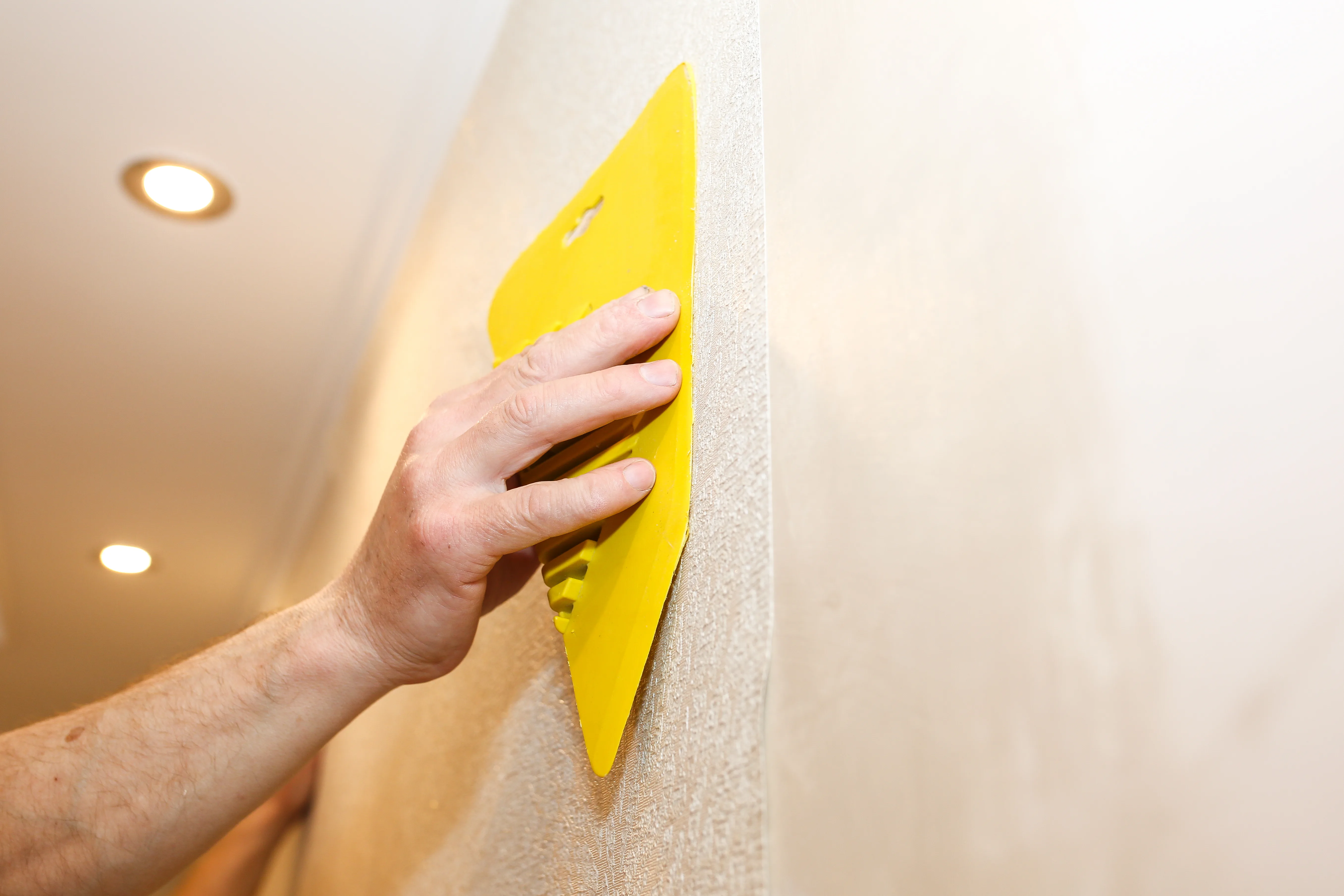 A hand using a yellow scraper on a textured wall during surface preparation