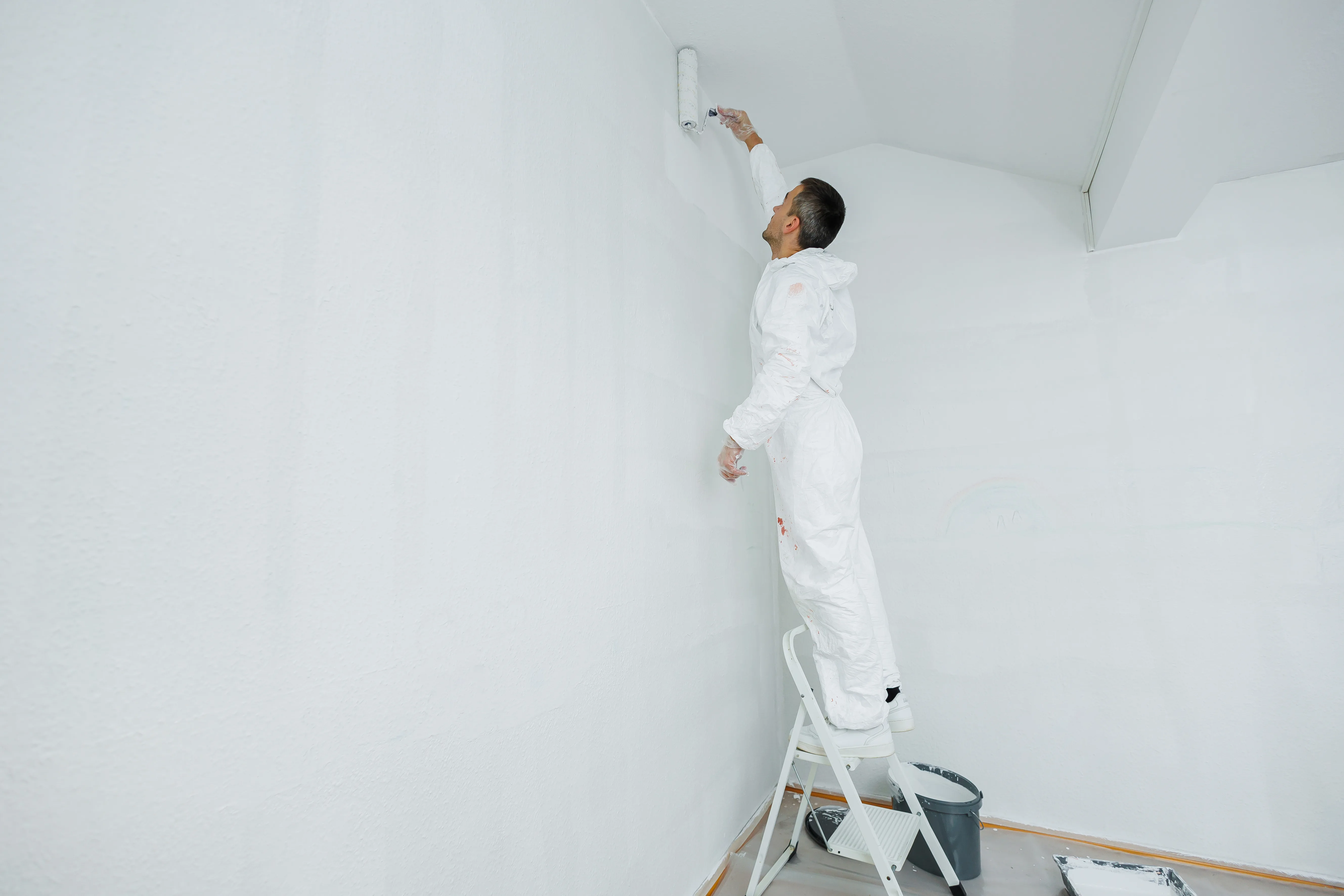 A painter in a white suit applying fresh paint to a wall using a roller on a ladder