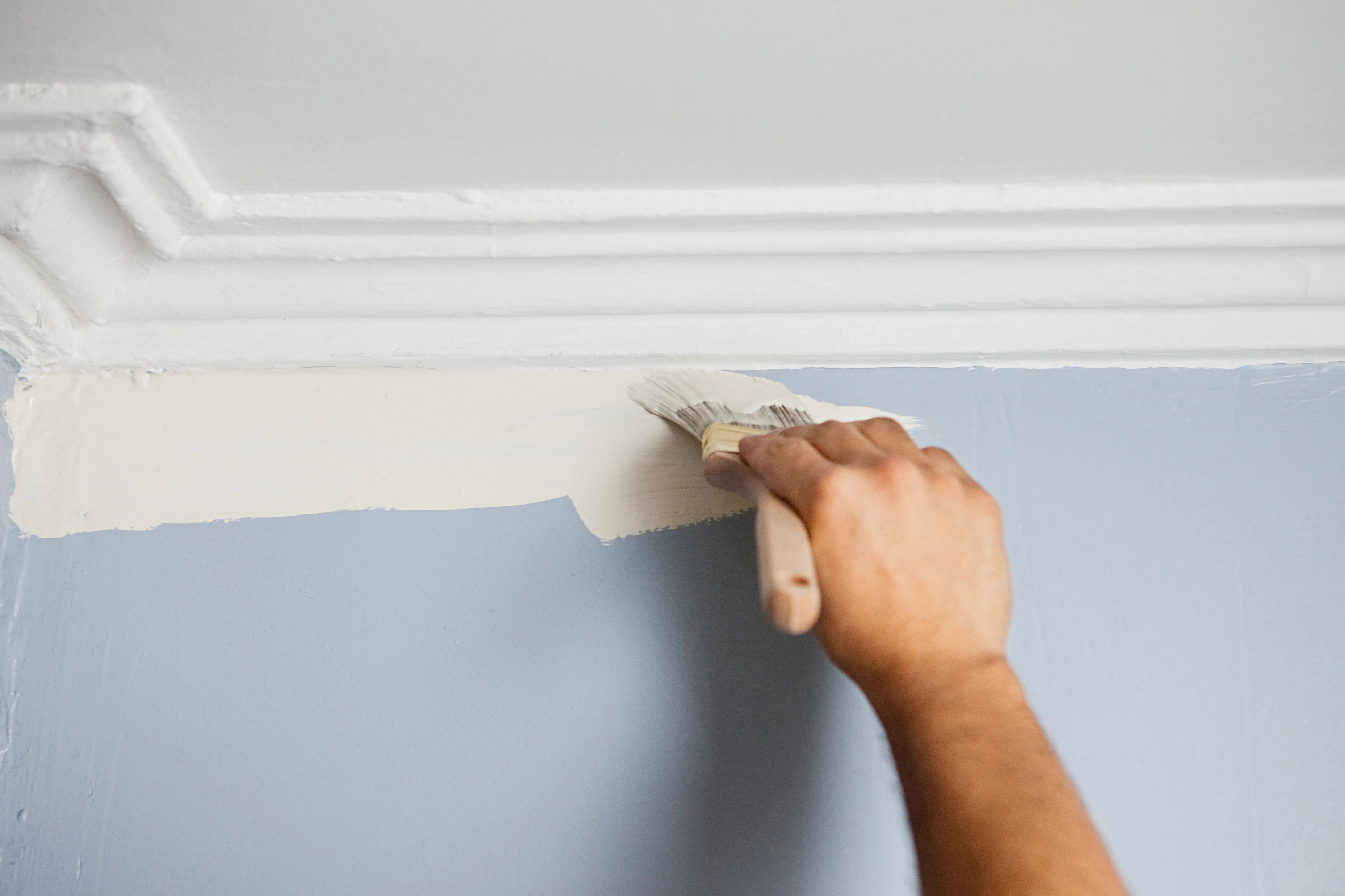 A hand applying paint to the top edge of a blue wall near the ceiling