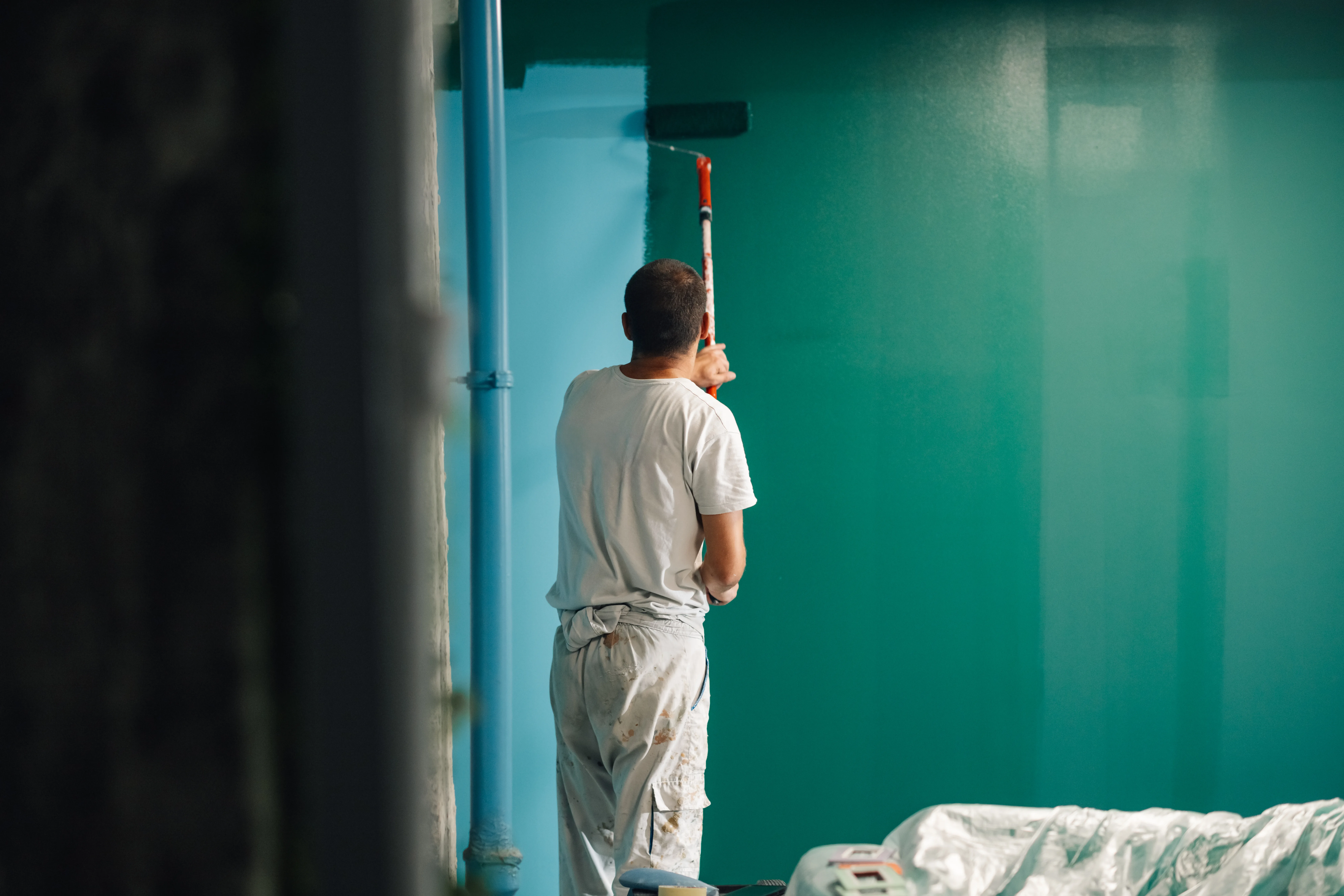 A painter applying teal paint on a wall using a roller in a well-lit interior space