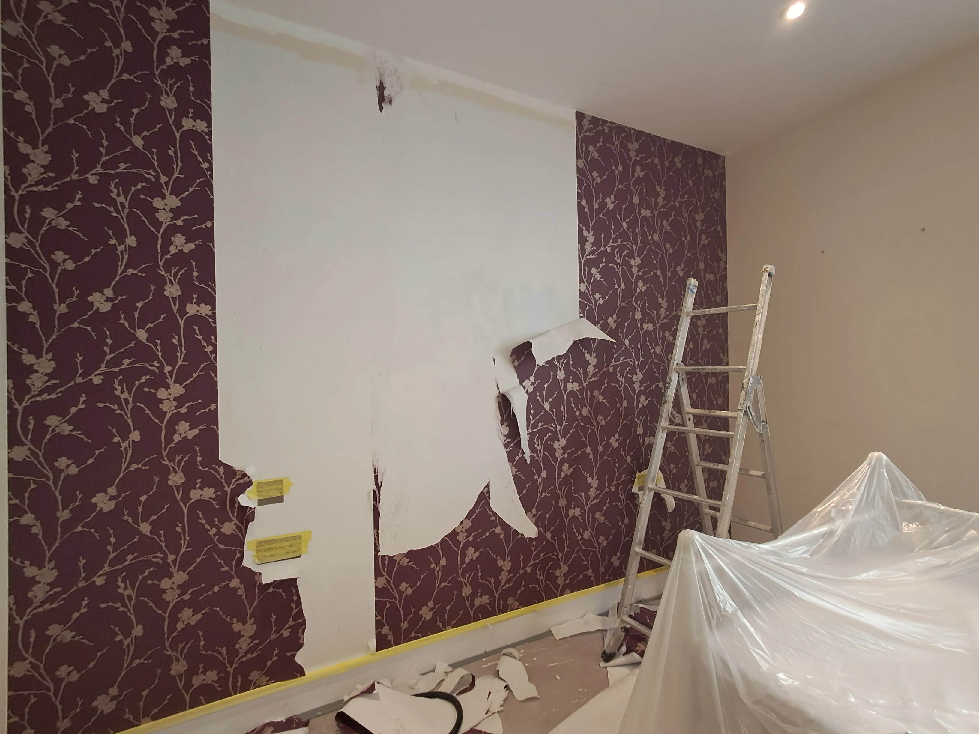 Wallpaper removal in progress with a stepladder and protective covering on the floor