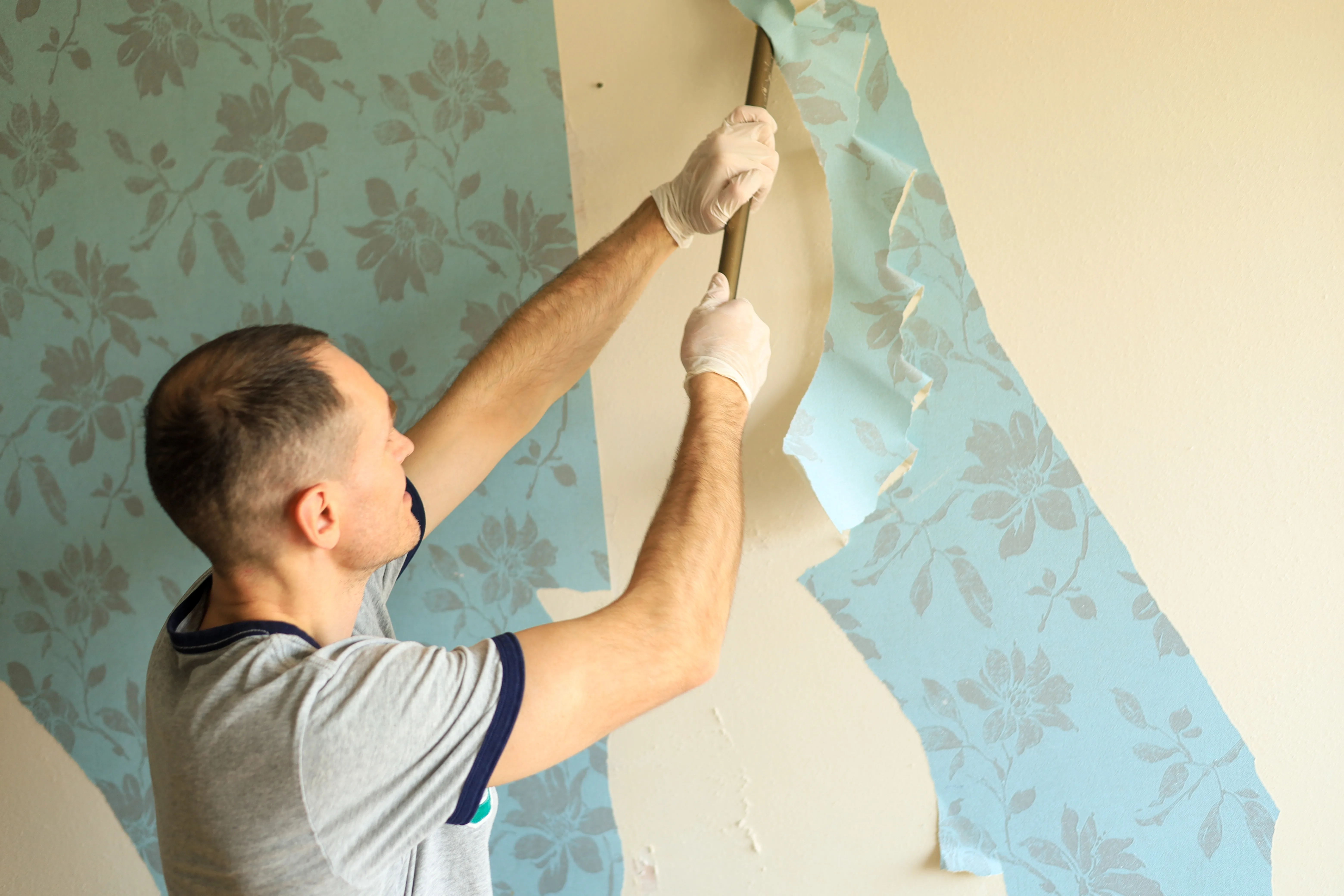 A person removing wallpaper from a wall using a scraper tool, wearing gloves