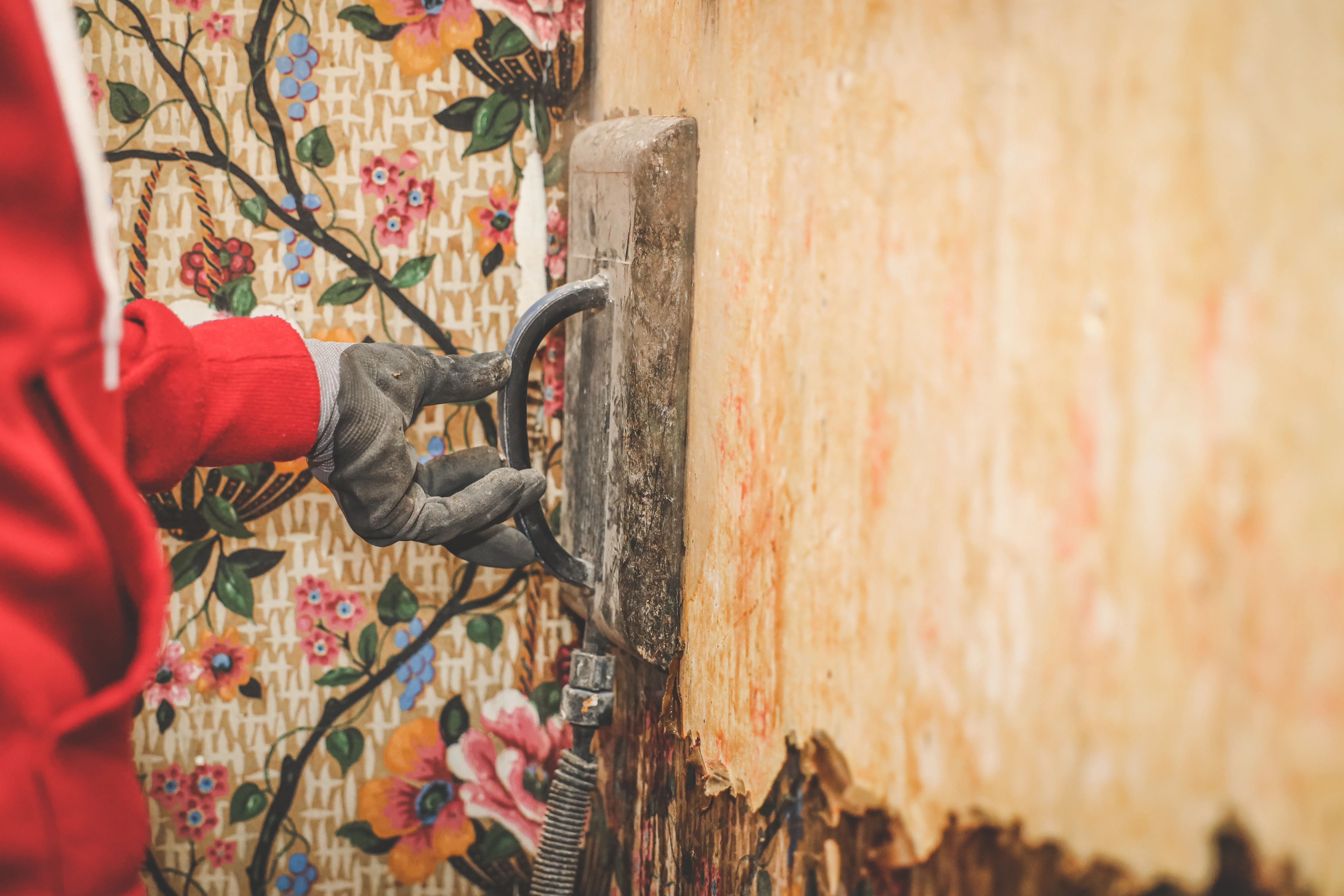 A worker using a trowel on a wall, preparing for wallpaper installation or removal, with floral patterned wallpaper visible